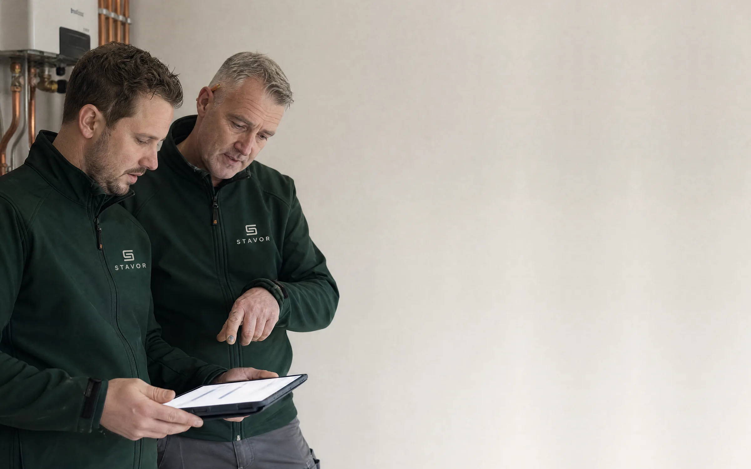 Twee monteurs in donkergroene Stavor-werkjassen kijken samen op een iPad, voor een cv-ketel met koperen leidingen